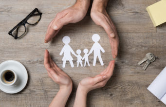 Close up of female and male hands protecting a paper chain family. Top view of two hands form a circle around white paper chain family on wooden table. Family care, insurance and helping hand concept.